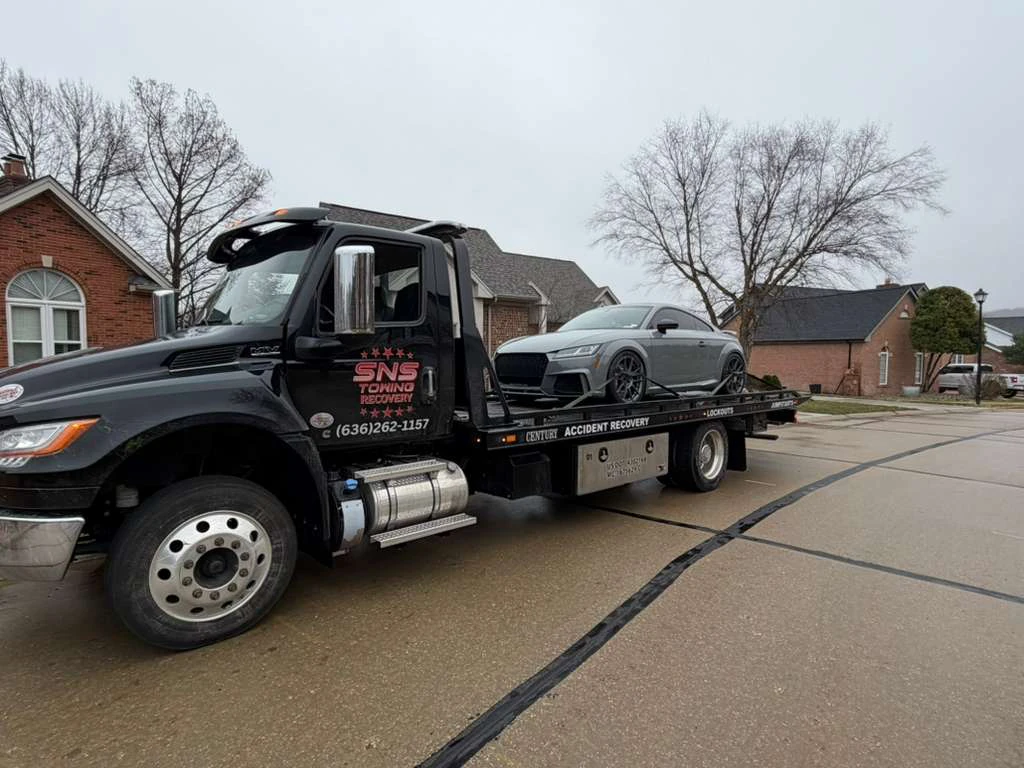 SNS Towing black flatbed truck carrying gray Audi sports car in residential neighborhood