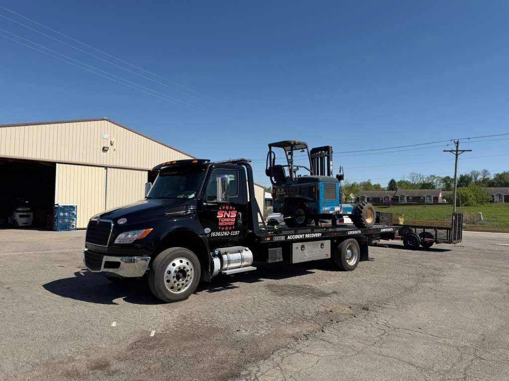 SNS Towing black flatbed truck carrying a blue mini excavator in a rural area