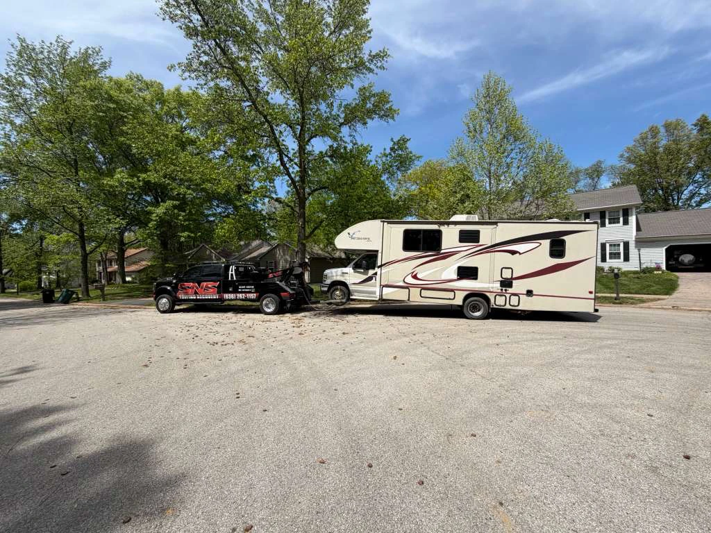 SNS Towing black wrecker truck towing a large RV motorhome in a residential neighborhood